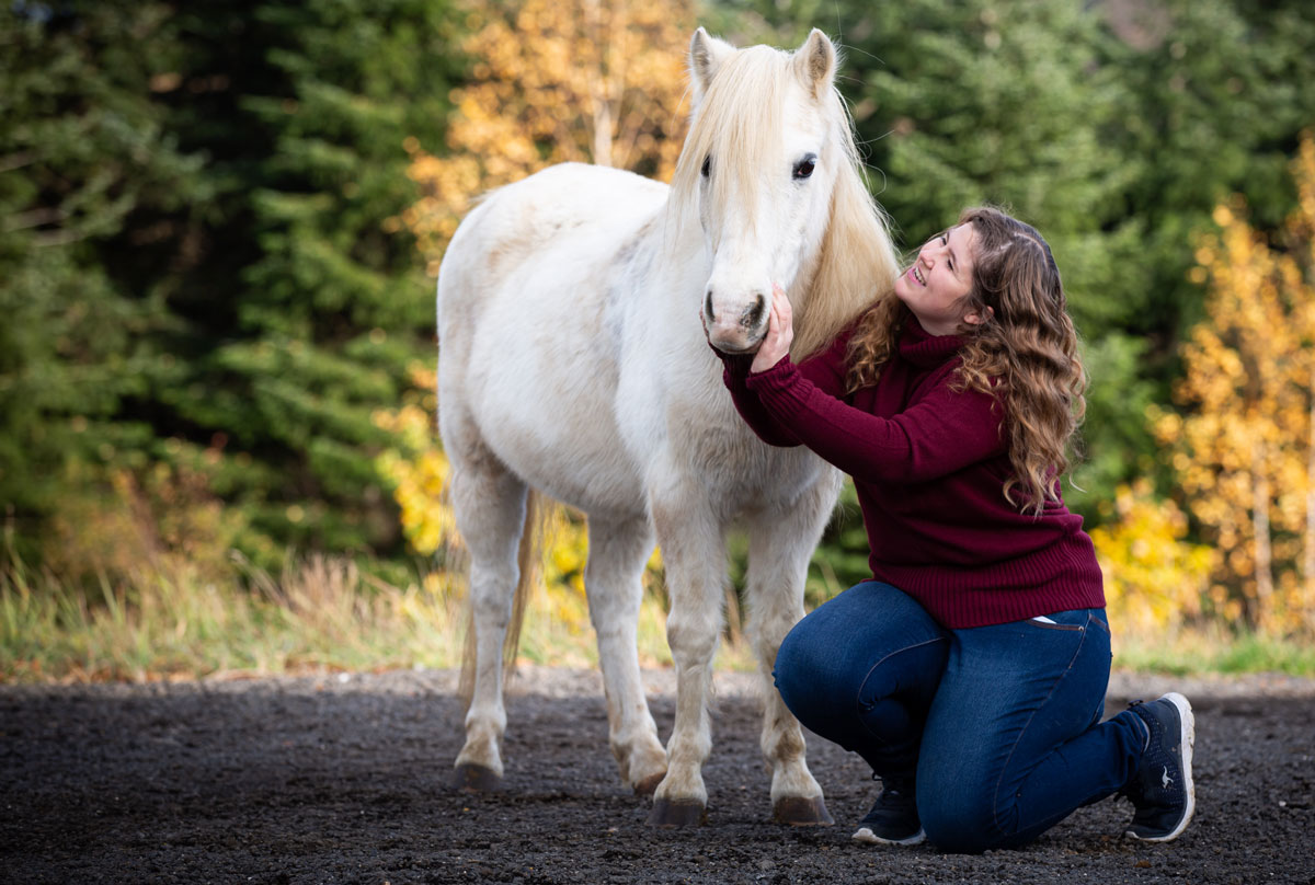 Lächelnde Frau mit lockigen Haaren kniet im Wald und streichelt liebevoll den Kopf eines weißen Pferdes.